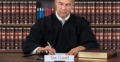 Judge with sign that says Tax Court; bookshelf behind him