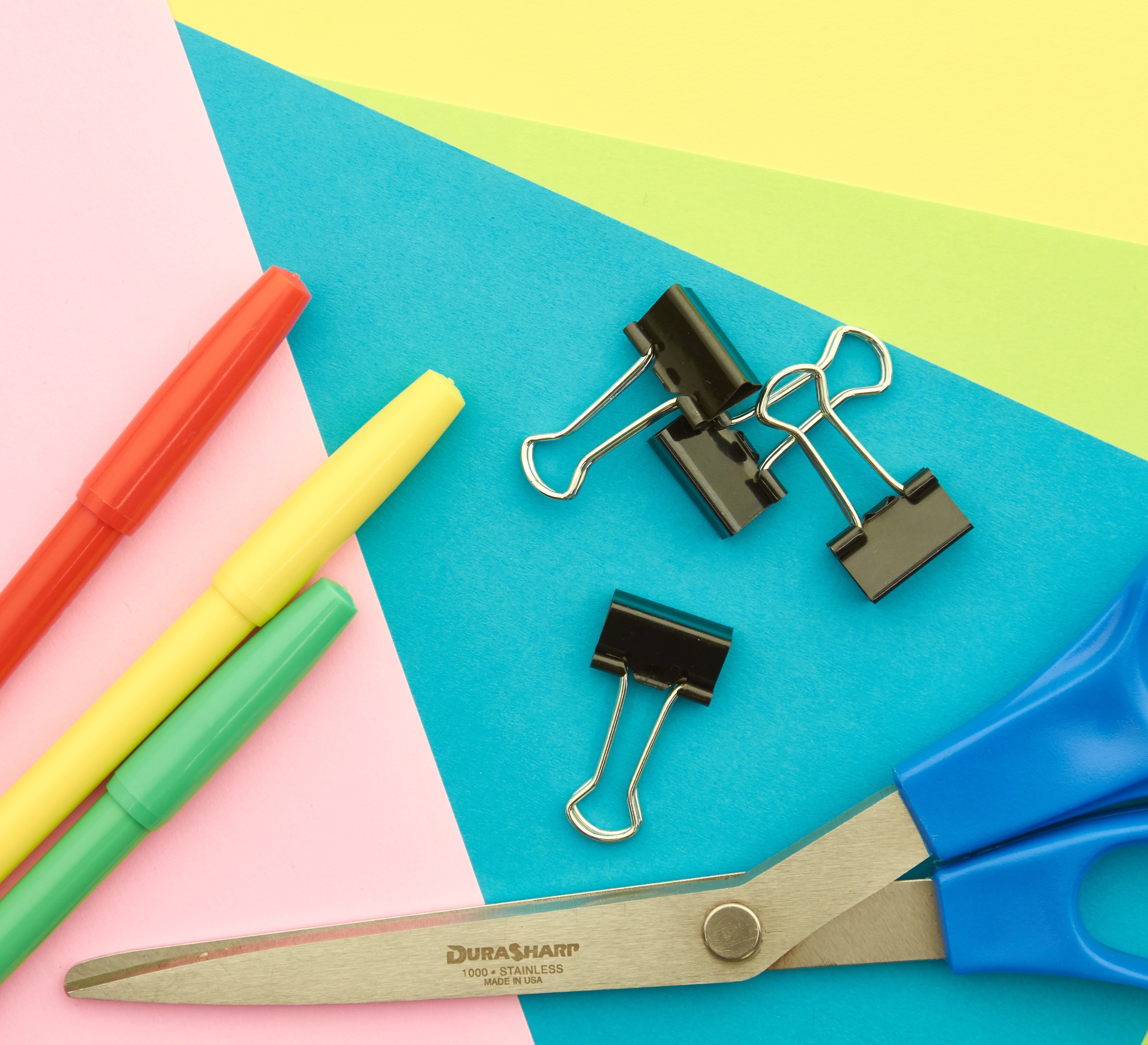 Colorful pens, binder clips, and scissors arranged on bright paper.