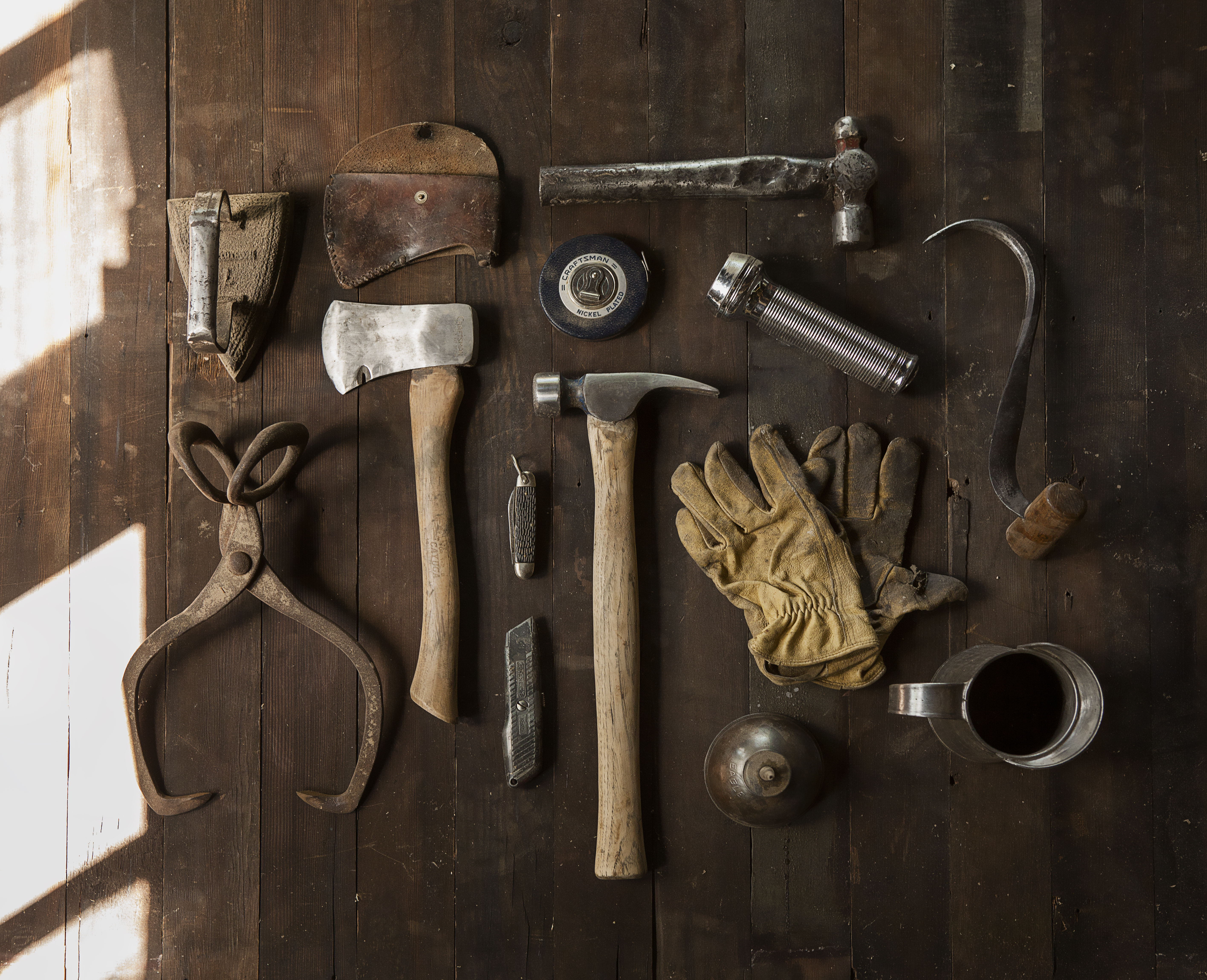 A collection of vintage hand tools, gloves, and a metal cup arranged on a wooden surface.